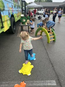 Children play on colorful outdoor equipment beside a brightly painted bus while adults gather under a canopy.