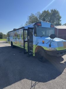 Light-blue and green school bus with an open door and colorful cartoon graphics, parked outside on a sunny day.